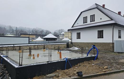 Plac budowy z wykonaną betonową płytą fundamentową i wystającym zbrojeniem, obok zabytkowego budynku w zimowej scenerii.