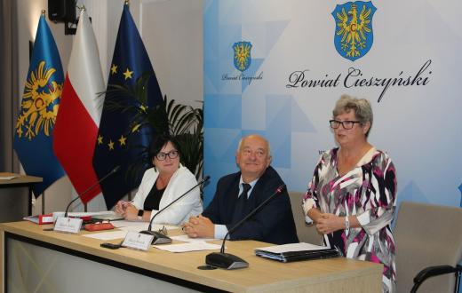 Uczestnicy VIII sesji Rady Seniorów Powiatu Cieszyńskiego przy stole obrad; kobieta przemawia, za nimi flagi i herb powiatu.