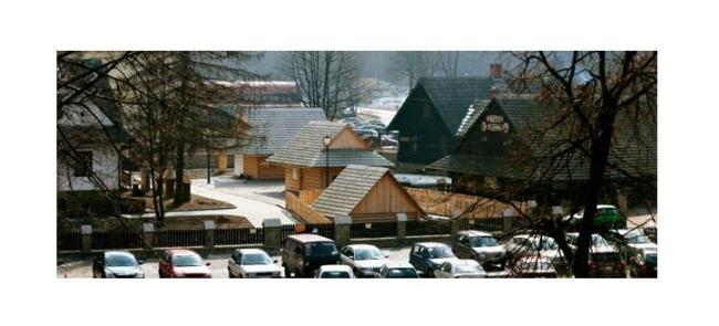 Drewniane budynki z dwuspadowymi dachami, otoczone ogrodzeniem. Na pierwszym planie zaparkowane samochody, w tle drzewa bez liści i inne zabudowania.