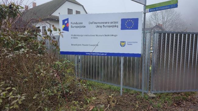 Metalowe ogrodzenie z zamontowaną tablicą informacyjną o dofinansowaniu z Unii Europejskiej. W tle budynek muzeum, krzewy i drzewa bez liści. Po prawej stronie widoczny fragment bramy i znak drogowy.